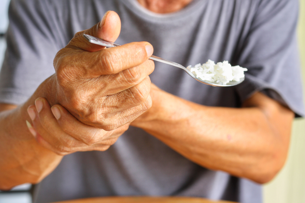 Elderly person struggling to eat due to hand tremors, holding spoon of rice
