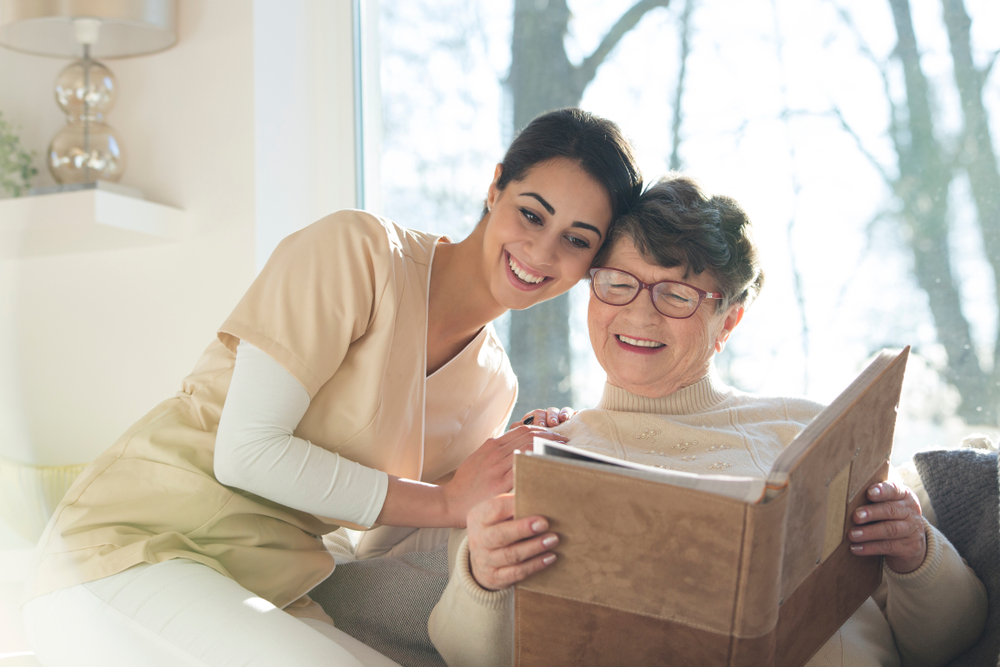 Home caregiver sharing joyful moment with elderly woman looking at photo album
