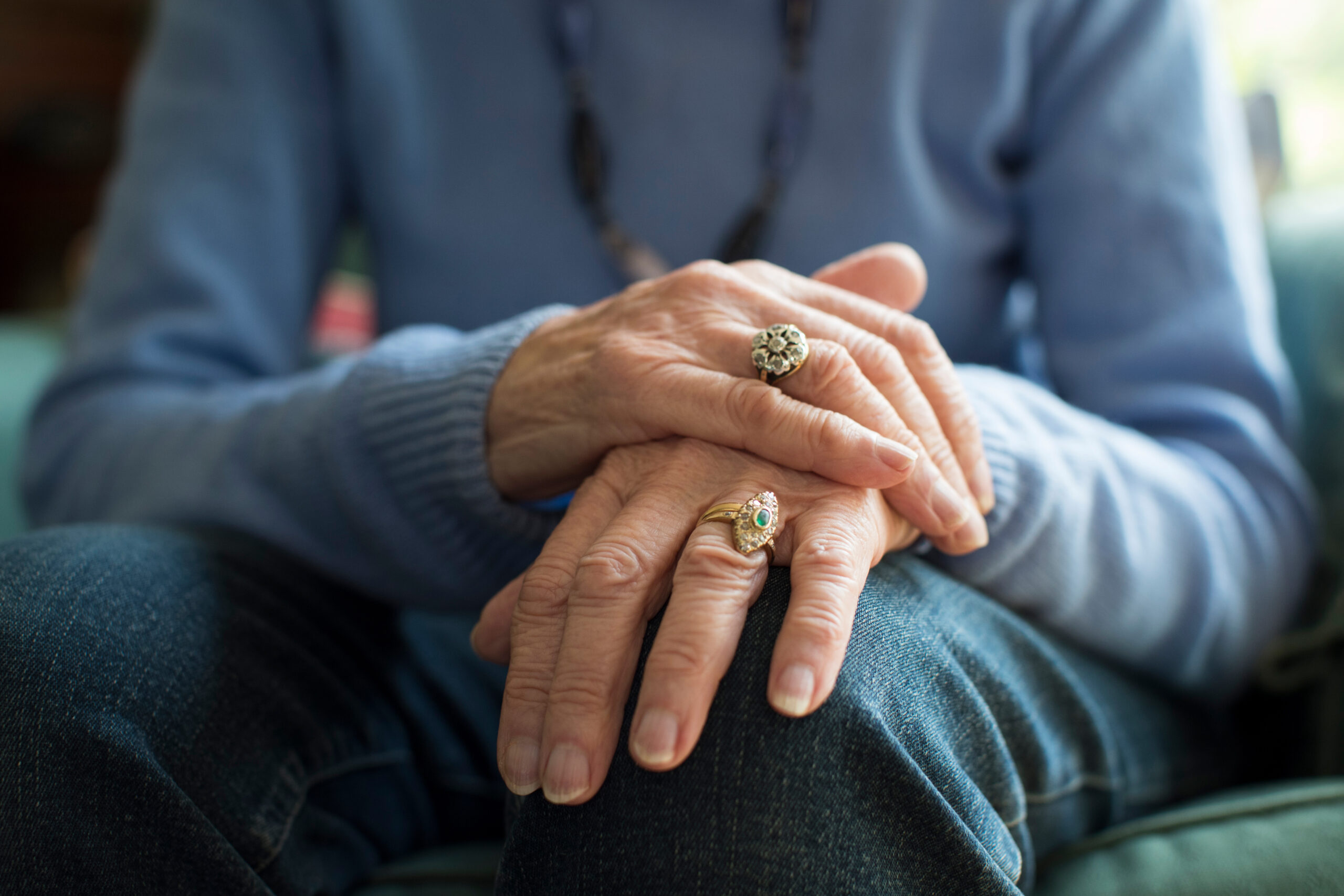 Close-up of an older person’s hands resting on their lap, wearing rings, with a focus on their fingers and blue clothing.