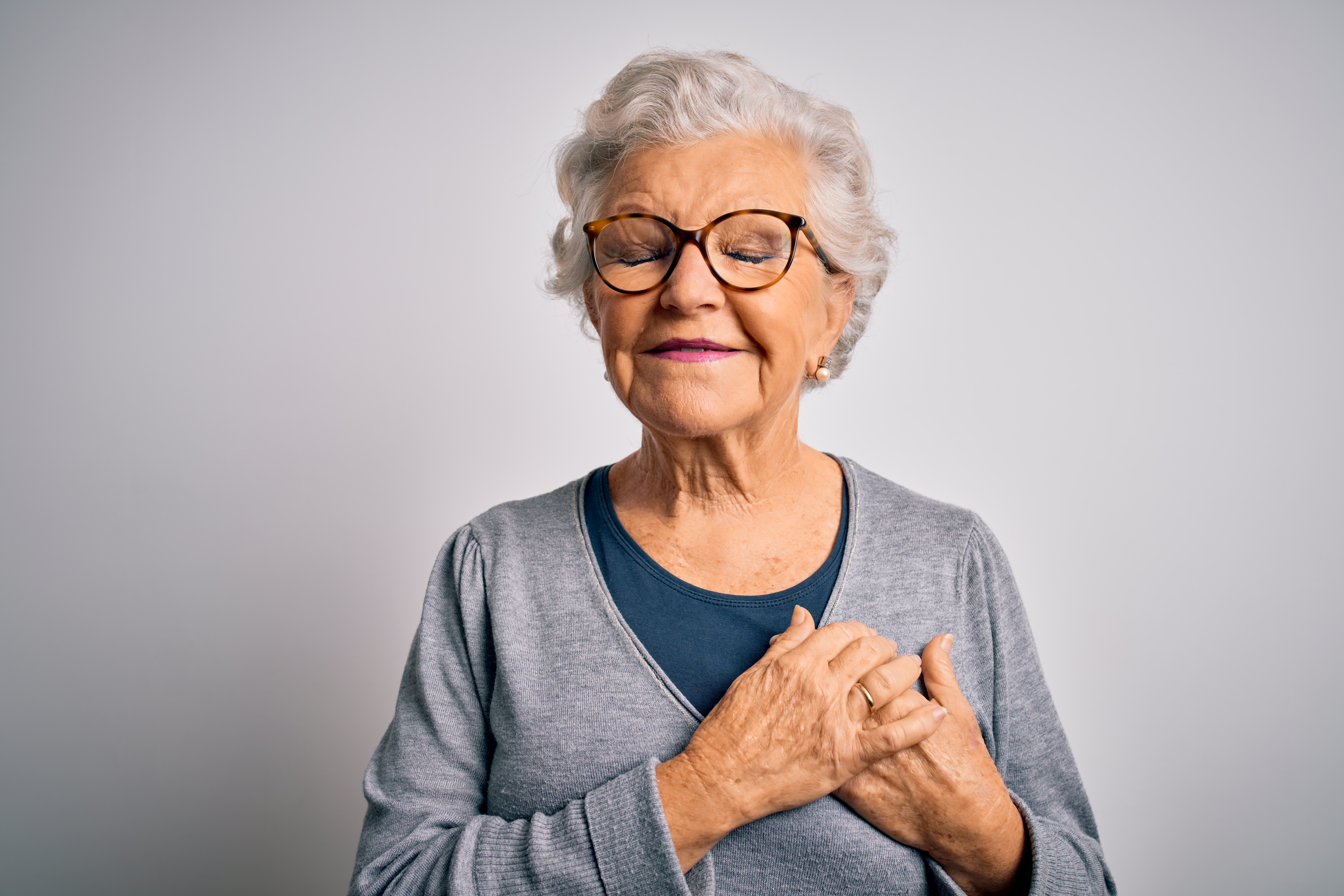 Older woman with short gray hair and glasses with her eyes closed, holding her hands over her chest and smiling gently.