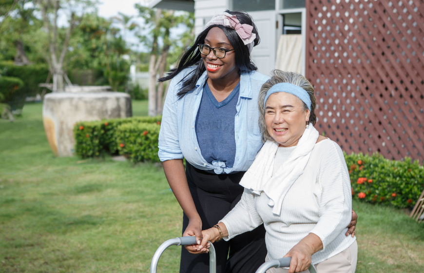 Senior Asian woman walking with walker and African American woman friend in garden showing home care supports independence.
