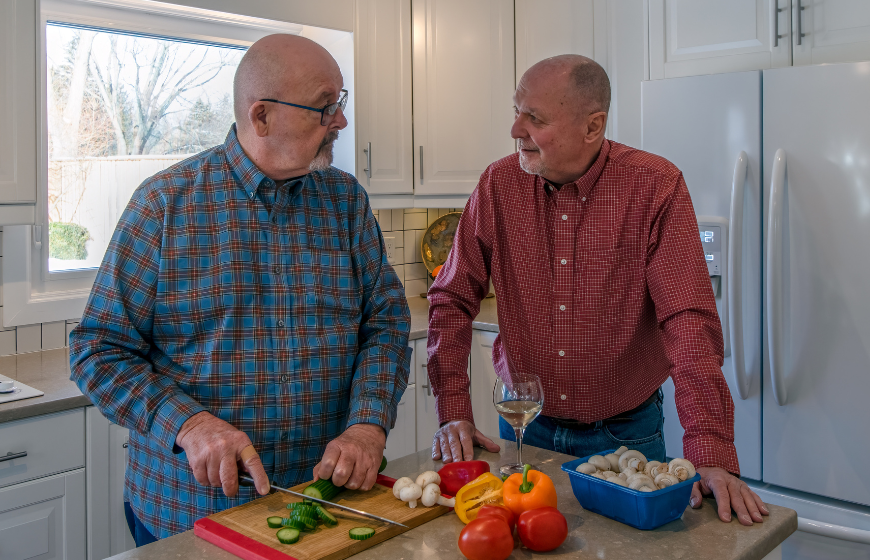 Couple of men engages in conversation about senior nutrition in their kitchen as one of them slices a cucumber and prepares food
